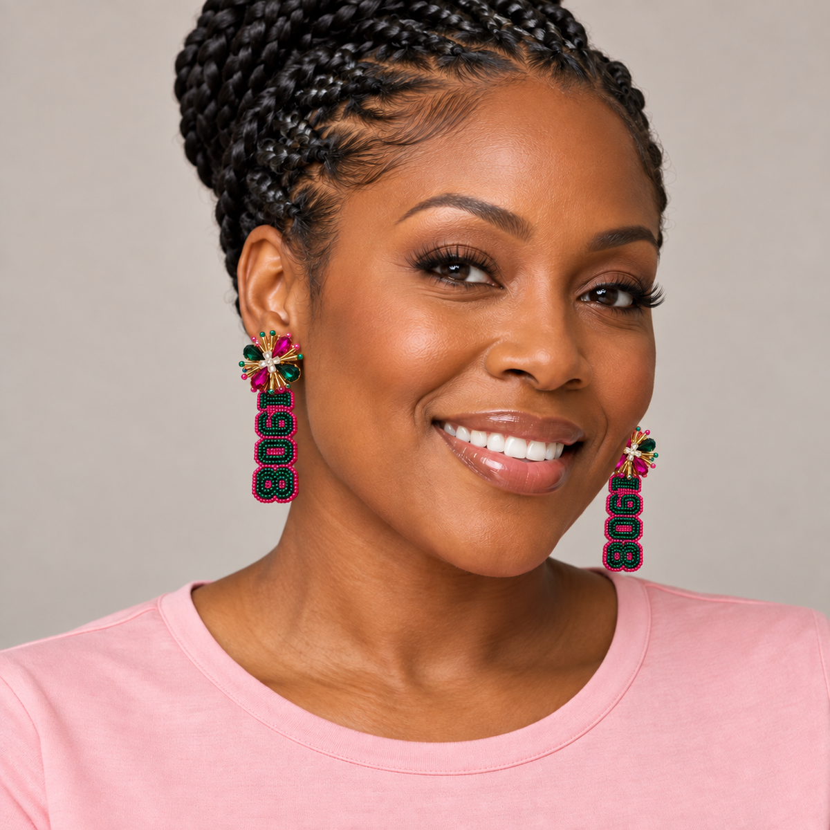 A smiling woman with braided hair wearing colorful dangling earrings with the numbers 1908, paired with a light pink top against a neutral gray background.