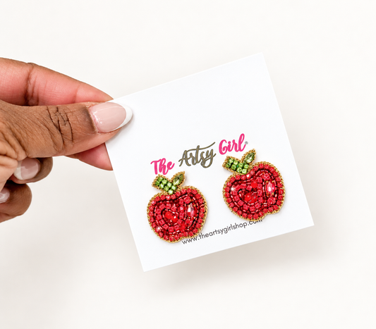 A hand holds a white card displaying two red apple-shaped stud earrings with green and gold beaded details. The card is branded 'The Artys Girl'.