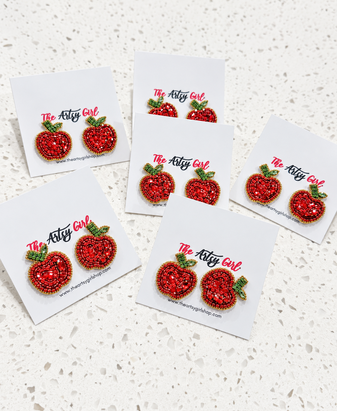 Five pairs of beaded apple stud earrings on white display cards, arranged on a light speckled surface. The earrings are red with green leaves and gold-toned outlines, and the cards show the brand name 'The Attic Girl'.