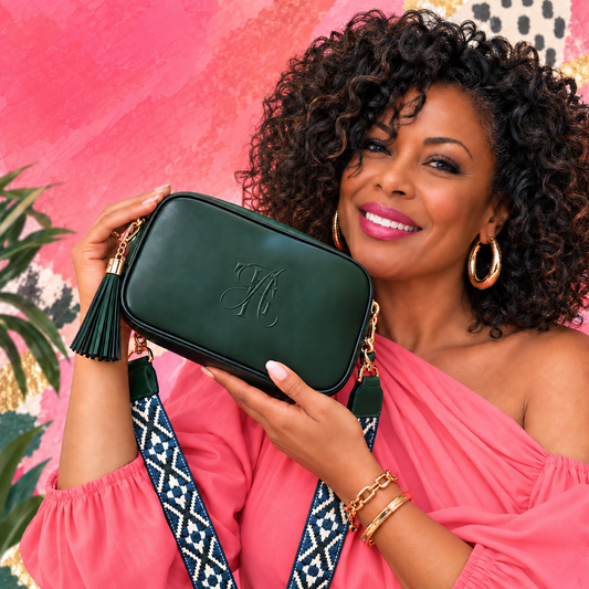 A woman in a coral pink off-the-shoulder top holds a black vegan-leather crossbody bag with a monogrammed The Artsy Girl logo, gold chain hardware, and a patterned blue and white strap against a vibrant pink background.