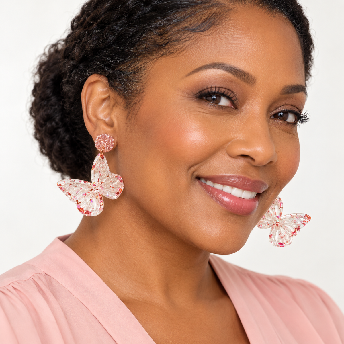 A close-up of a woman wearing large butterfly-shaped drop earrings with pink rhinestone detailing and a textured circular stud, paired with a light pink top against a clean white background.