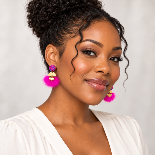A close-up portrait of a woman wearing bright pink fluffy tassel earrings with gold circular accents and purple floral tops, paired with a white blouse against a light gray background.
