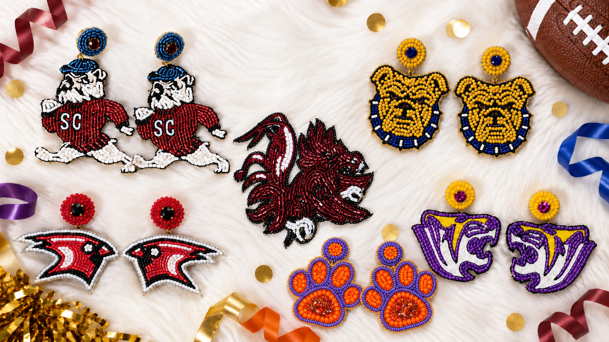 A flat-lay display of multiple embroidered collegiate bulldog and mascot earrings on a white faux fur background with gold confetti, ribbons, and a football prop. The earrings include designs for the South Carolina Gamecocks, Clemson Tigers, Cardinal Newman, Benedict Tigers, NCA&T, and SC State Bulldogs.
