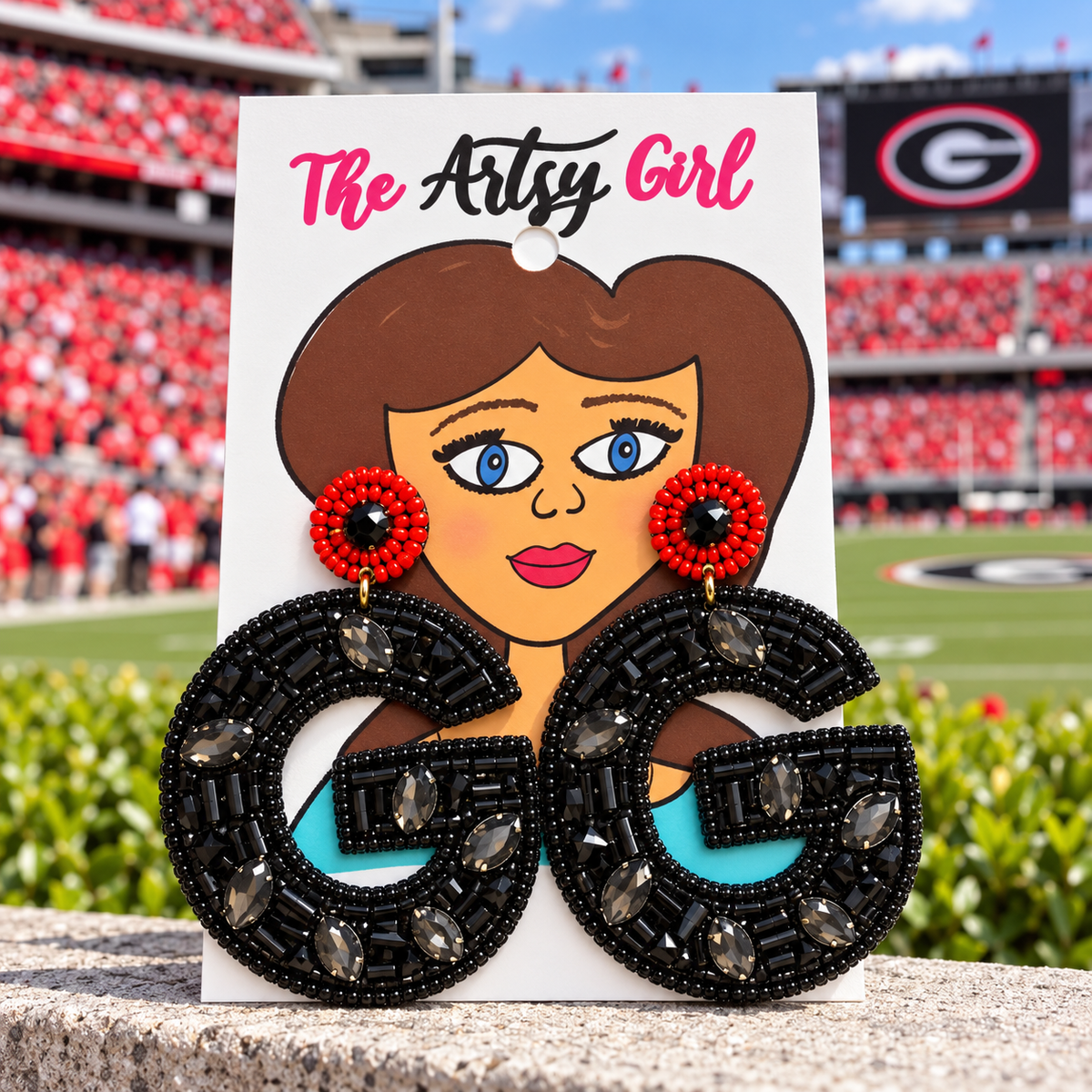 A pair of large letter G drop earrings displayed on a white card printed with a cartoon face and the text 'The Artsy Girl'. The earrings feature black beaded letter Gs with clear rhinestones, red beaded circular studs, and turquoise blue accents.