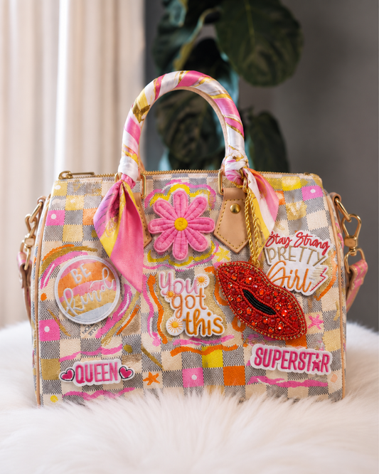 A highly decorated, patchwork-style handbag with pink and gold checkered patterns, embellished with a pink satin scarf, a pink flower applique, a red sequined lip bag charm, and various text patches including 'Queen', 'Superstar', and 'Be Lovely'.