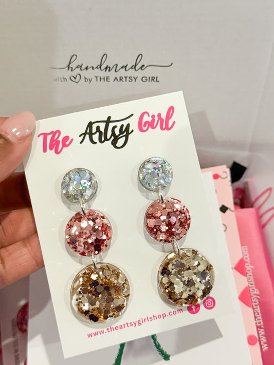 A pair of metallic earrings with rose gold, silver, and gold colors, displayed on a card with 'The Artsy Girl' branding.