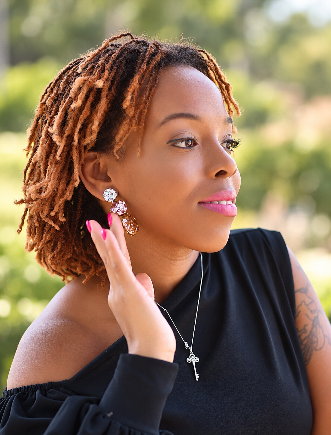 A woman with short brown dreadlocks is shown in a close-up outdoor portrait, wearing a black off-the-shoulder top, a silver key necklace, and a dangling earring with clear stones and gold-toned details.