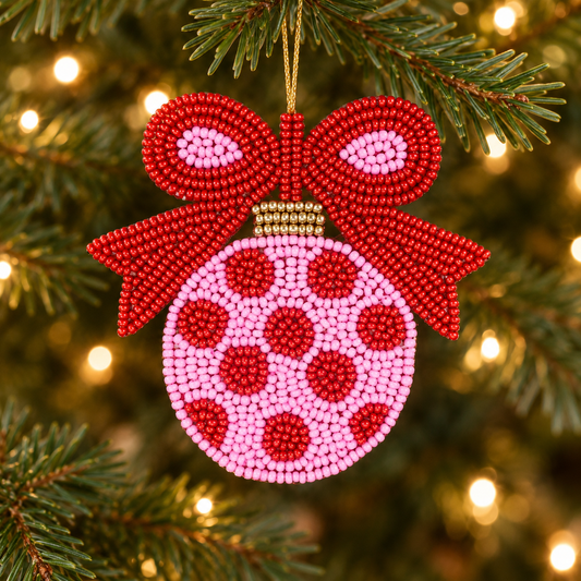 Pink and red beaded Christmas ornament with a large red bow hanging on an evergreen tree with warm white lights.