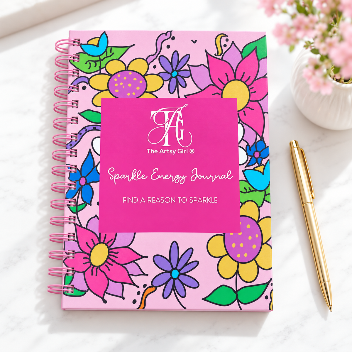 Floral Sparkle Energy Journal on a white desk beside a gold pen and pink flowers, with “Find a Reason to Sparkle” on the cover.