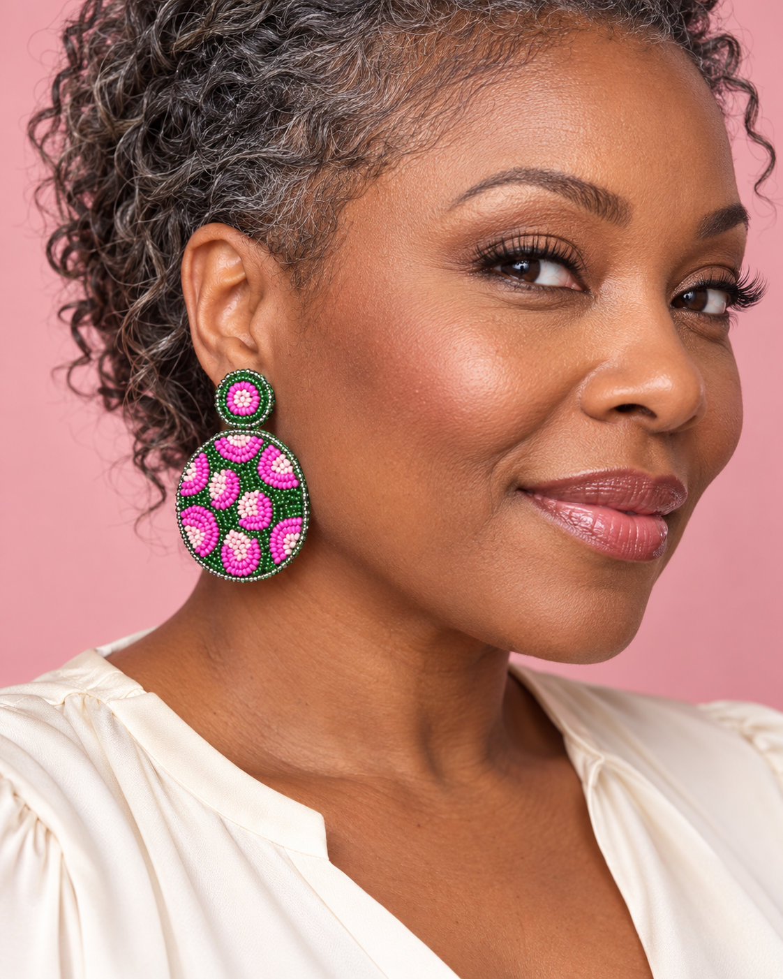 Woman wearing round pink and green beaded leopard earrings with a cream blouse against a blush pink background.
