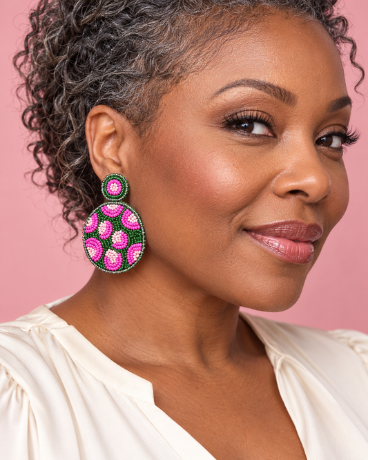 Woman wearing round pink and green beaded leopard earrings with a cream blouse against a blush pink background.