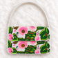 A small structured handbag with a white handle and body, covered in a colorful beaded floral pattern of pink flowers and green leaves on a white background, laid flat on a white faux fur surface.