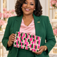 A woman wearing a green blazer and matching pants over a pink satin top, holding a patterned clutch bag with pink, green, and beige brushstroke motifs.