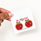 A hand holds a white card displaying two red apple-shaped stud earrings with green and gold beaded details. The card is branded 'The Artys Girl'.