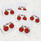 Five pairs of beaded apple stud earrings on white display cards, arranged on a light speckled surface. The earrings are red with green leaves and gold-toned outlines, and the cards show the brand name 'The Attic Girl'.