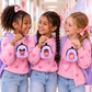 Three young girls standing together in a school hallway, each holding a small purple and pink backpack-style pouch with cartoon character graphics. They are wearing pink heart-patterned sweaters, blue jeans, and purple backpacks.