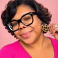 A close-up portrait of a woman with short curly black hair, wearing thick black cat-eye glasses with small gold stars and a bright pink top. She is holding up a round leopard-print earring against a solid pink background.