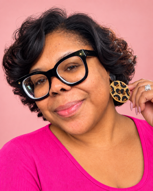 A close-up portrait of a woman with short curly black hair, wearing thick black cat-eye glasses with small gold stars and a bright pink top. She is holding up a round leopard-print earring against a solid pink background.