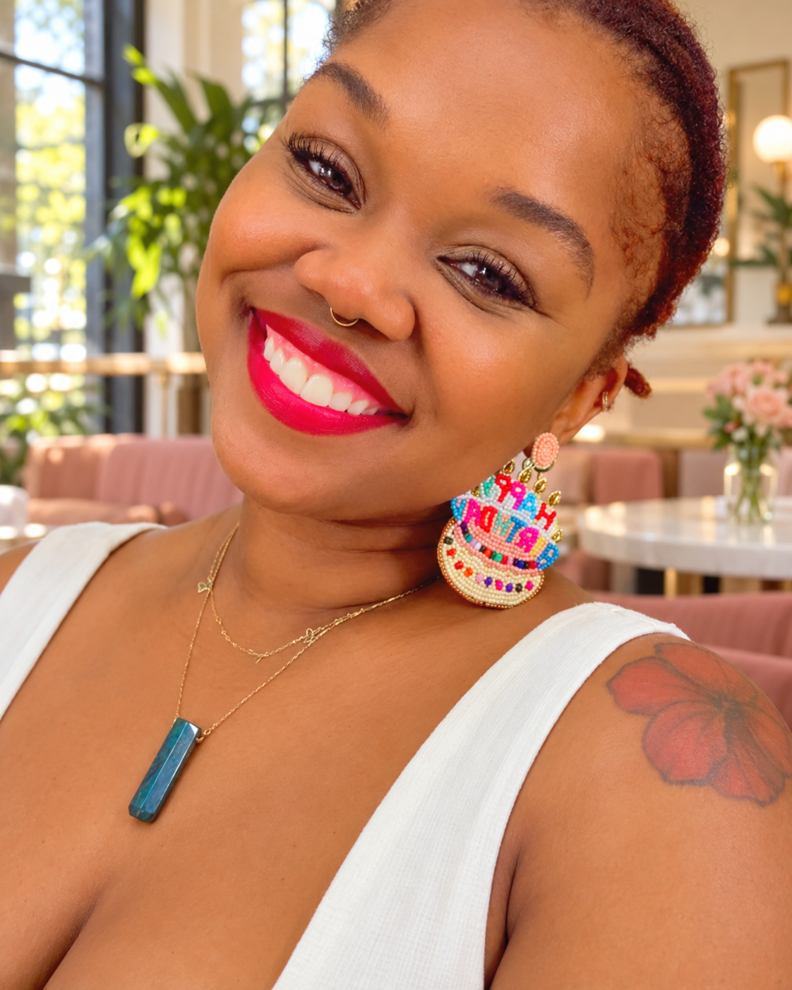 A smiling woman wearing a colorful, tiered shell-shaped earring with pink, blue, yellow, and white details, along with a gold necklace and white top, in a bright indoor setting.
