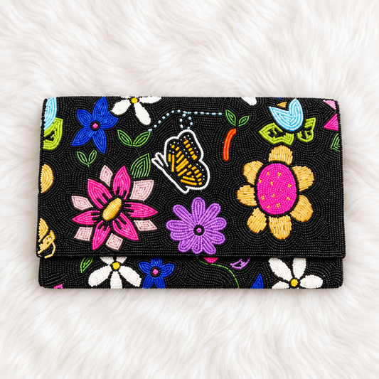 A black beaded clutch wallet featuring a vibrant, multicolor floral and butterfly pattern, laid flat on a white faux fur background.