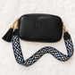 A black vegan-leather crossbody bag with an embossed monogram logo, featuring a black tassel charm and a navy blue and white geometric patterned woven strap with gold-tone hardware, laid flat on a white faux fur background.