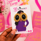 A hand holds a white card displaying a pair of statement earrings, with large black oval eyes and gold textured cheeks. The background is a pink-themed desk with notebooks and decorative items.