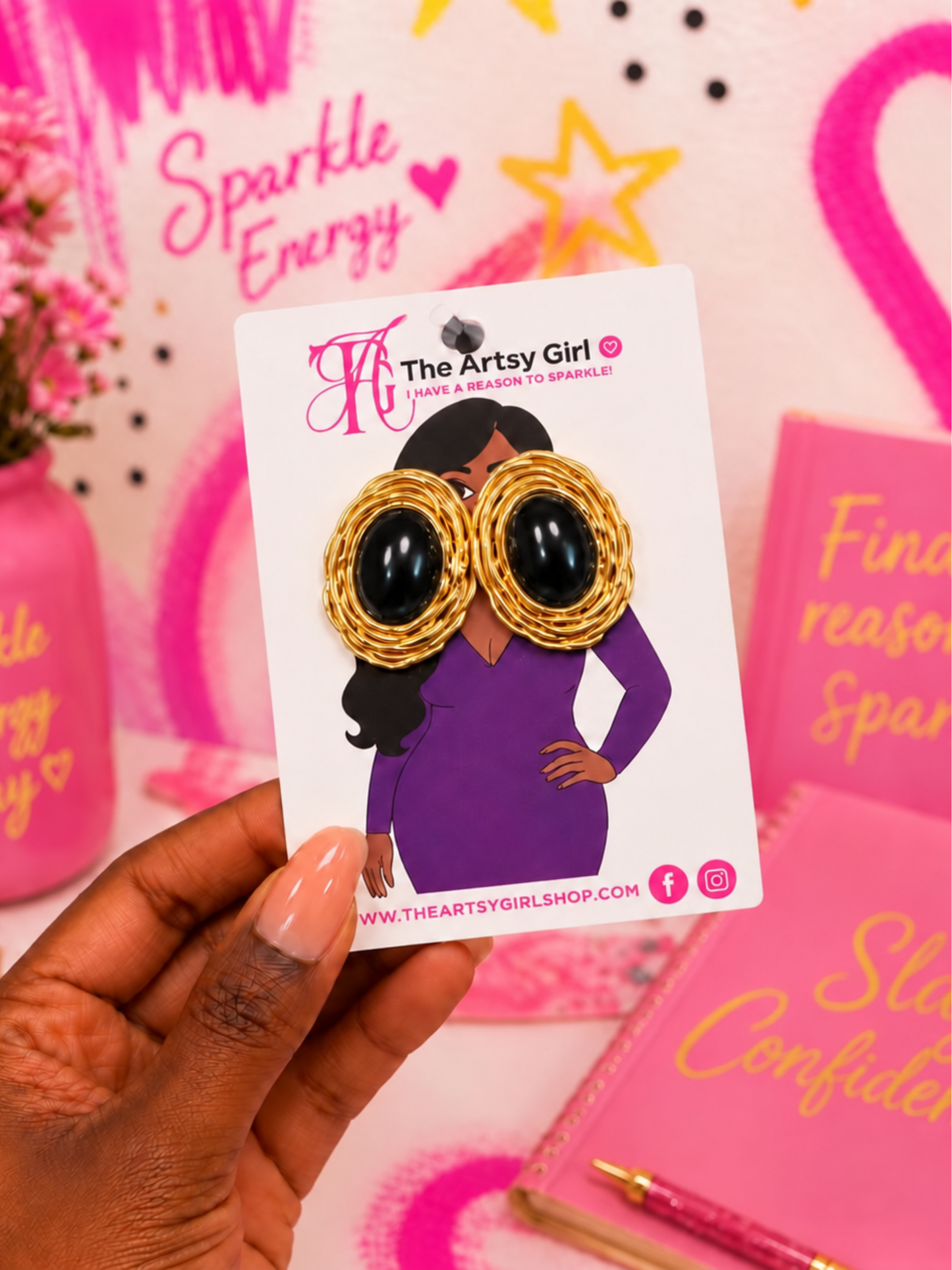 A hand holds a white card displaying a pair of statement earrings, with large black oval eyes and gold textured cheeks. The background is a pink-themed desk with notebooks and decorative items.