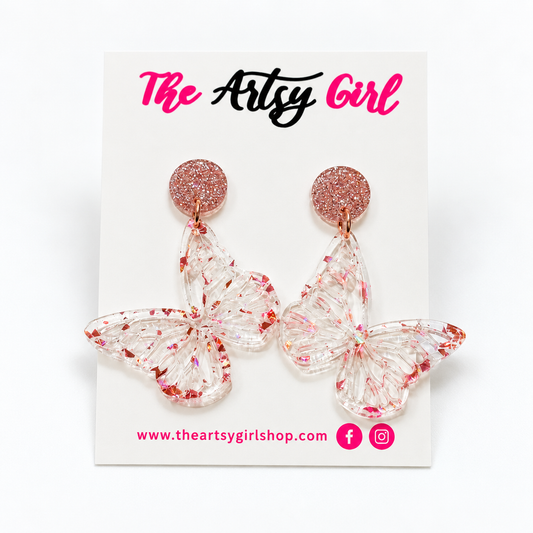 A pair of butterfly-shaped dangling earrings with glittery rose gold circular studs and clear wings filled with red glitter, displayed on a white card labeled 'The Artsy Girl'.