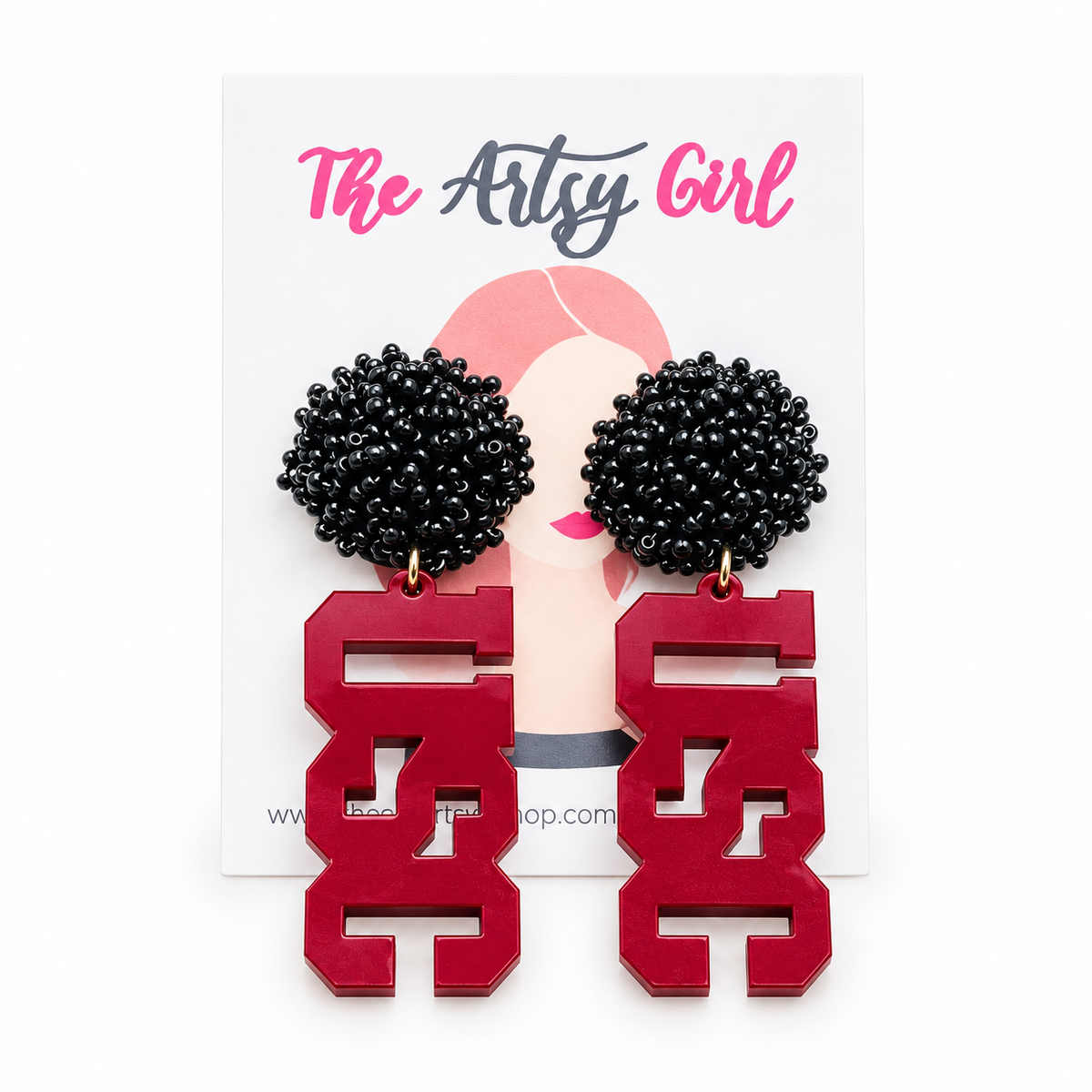 A pair of statement earrings displayed on a white card from 'The Artsy Girl'. The earrings feature black beaded pom-poms on the top and garnet acrylic letters on the bottom, suspended by small gold rings.
