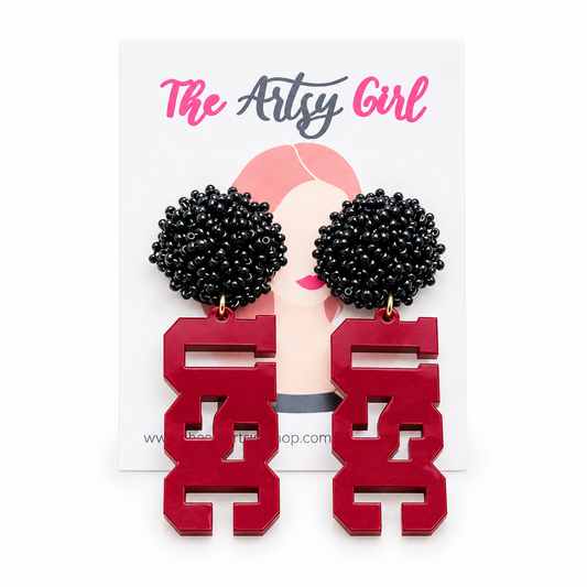 A pair of statement earrings displayed on a white card from 'The Artsy Girl'. The earrings feature black beaded pom-poms on the top and garnet acrylic letters on the bottom, suspended by small gold rings.