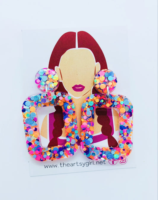 A pair of multicolored round square earrings with a glittery finish, featuring a design that resembles a woman's profile with earrings and a name tag.