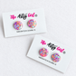 Two round stud earrings covered in multicolor glitter, displayed on white cards under the brand name 'The Artsy Girl'.