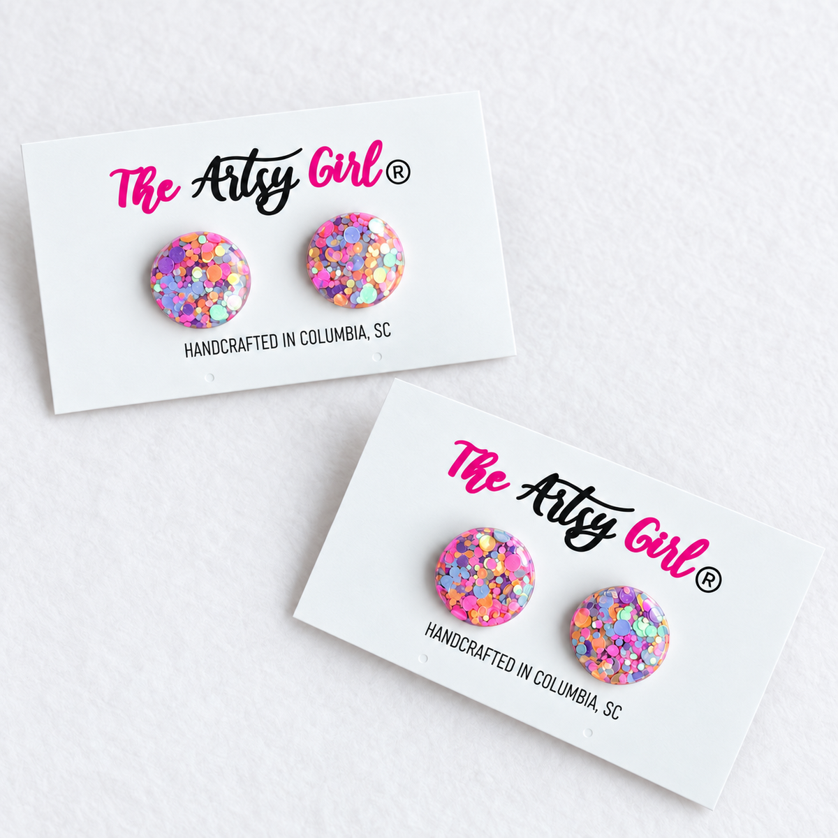 Two round stud earrings covered in multicolor glitter, displayed on white cards under the brand name 'The Artsy Girl'.