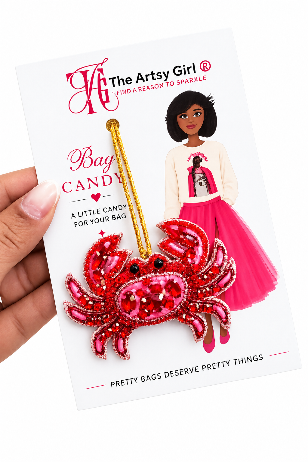 A hand holds a white retail card featuring a red beaded crab-shaped bag charm with a gold hanging loop. The card has the brand name 'The Artsy Girl' and includes an illustration of a woman in a pink outfit.