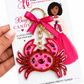 A hand holds a packaged keychain featuring a pink and red beaded crab charm with a bright pink ribbon and gold key ring, attached to a white display card from The Arts Girl.
