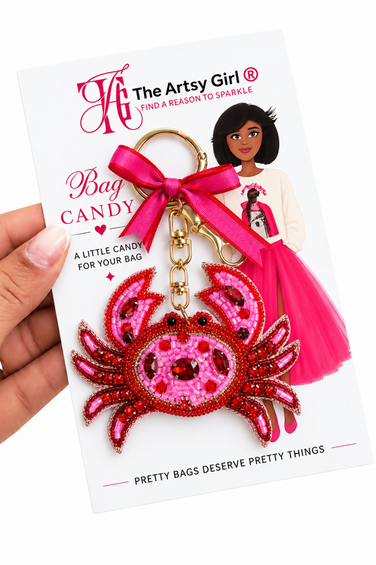A hand holds a packaged keychain featuring a pink and red beaded crab charm with a bright pink ribbon and gold key ring, attached to a white display card from The Arts Girl.