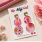 Pink and gold earrings on a 'The Artsy Girl' branded card with flowers in the background.