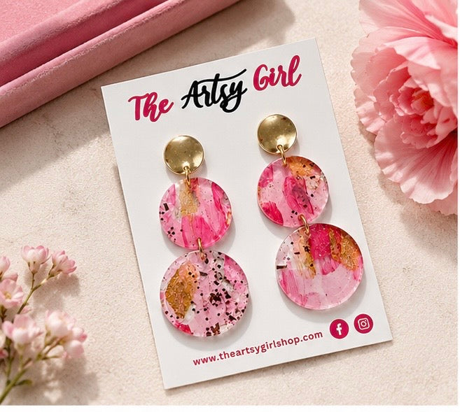 Pink and gold earrings on a 'The Artsy Girl' branded card with flowers in the background.