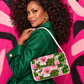 A woman with voluminous dark curly hair is shown from the back wearing a bright green satin jacket and holding a structured flap shoulder bag. The bag features a vibrant pink and green floral pattern with white trim and a white shoulder strap.