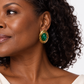 A close-up of a woman wearing a gold-toned drop earring with a green oval stone, shown against a clean white background.