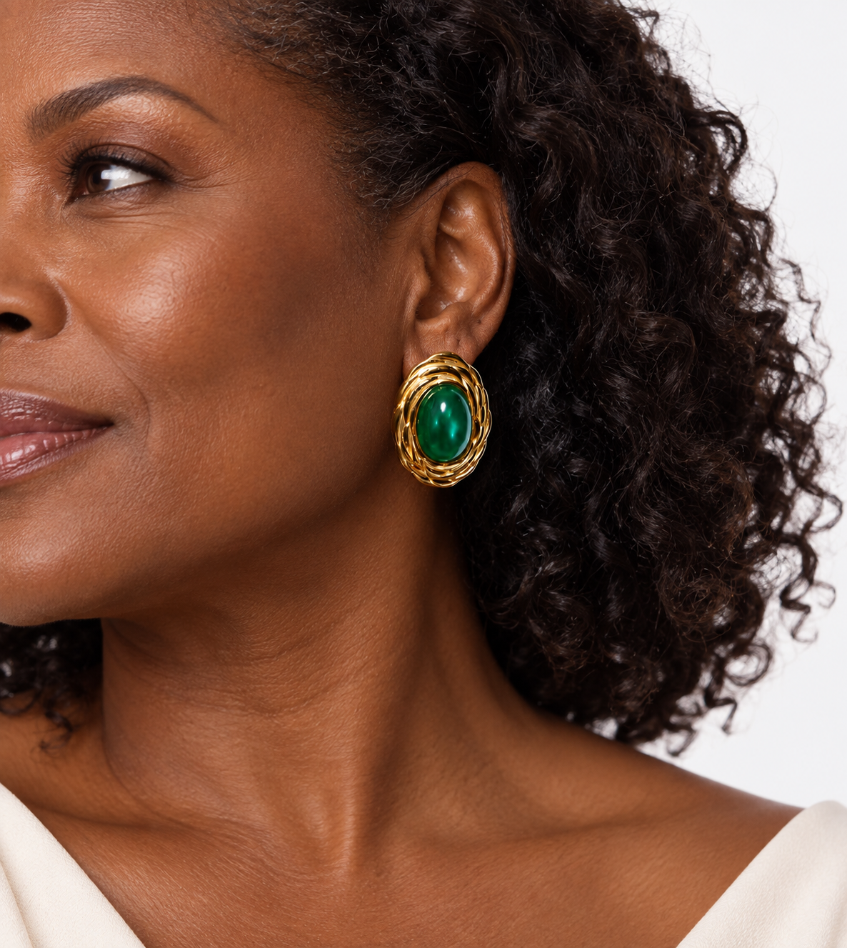 A close-up of a woman wearing a gold-toned drop earring with a green oval stone, shown against a clean white background.
