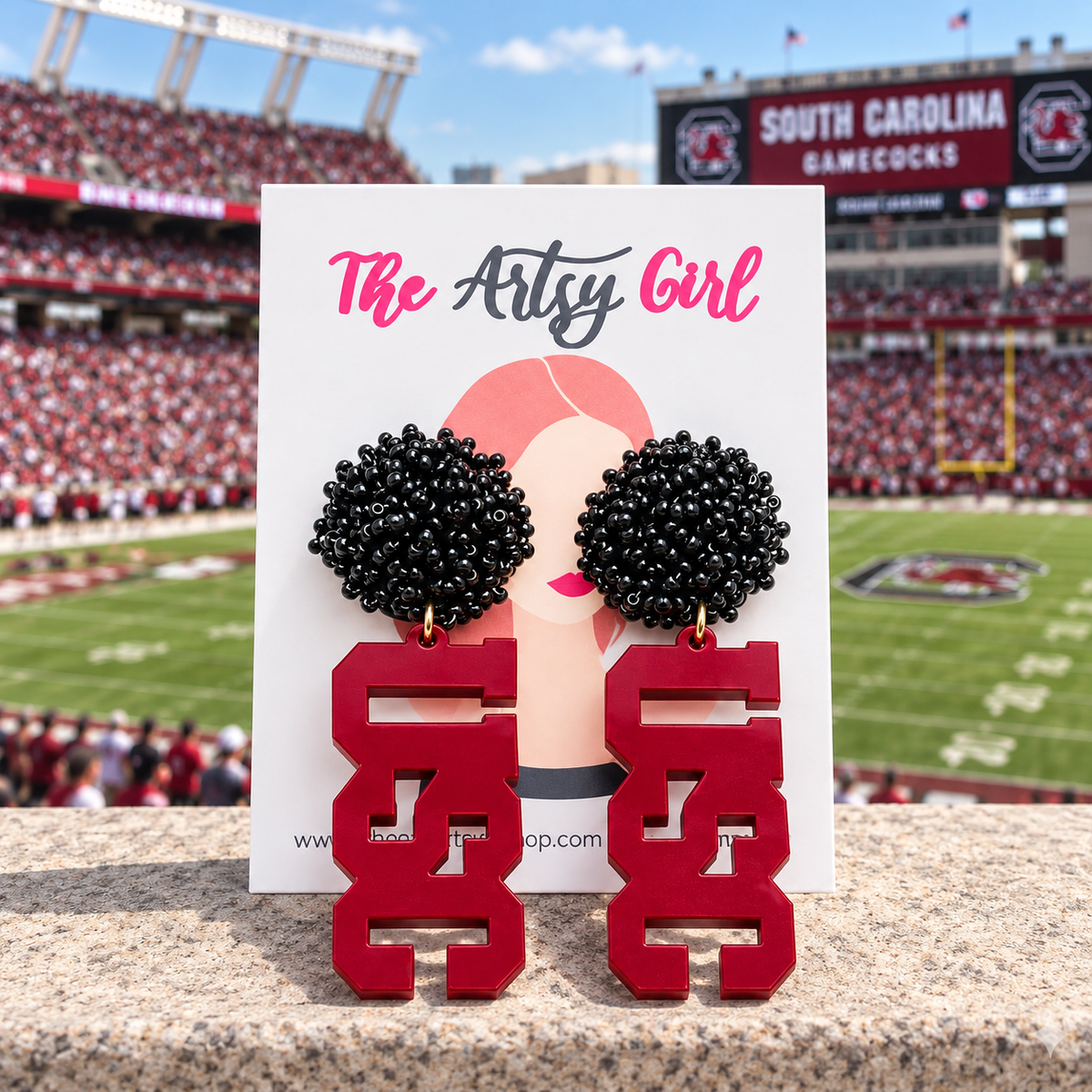 A pair of red and black South Carolina Gamecocks dangle earrings displayed on a white card that reads 'The Artsy Girl'. The background shows a blurred football stadium with fans and the South Carolina Gamecocks branding.