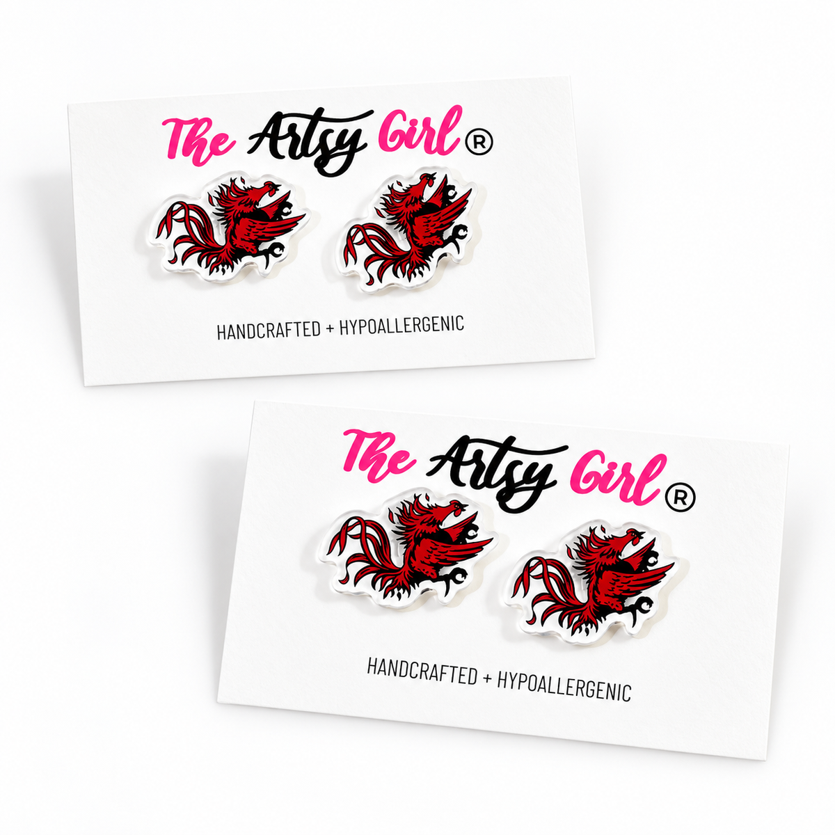 Two white display cards showing the same pair of Gamecock inspired stud earrings from different angles on a clean white background. The cards are branded 'The Artys Girl' in pink and black script, with 'HANDCRAFTED + HYOALLERGGENIC' printed below.
