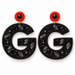 A pair of statement earrings featuring a large black beaded letter 'G' adorned with dark gray faceted marquise and oval crystals, topped with a red beaded circle and a central black faceted gemstone.