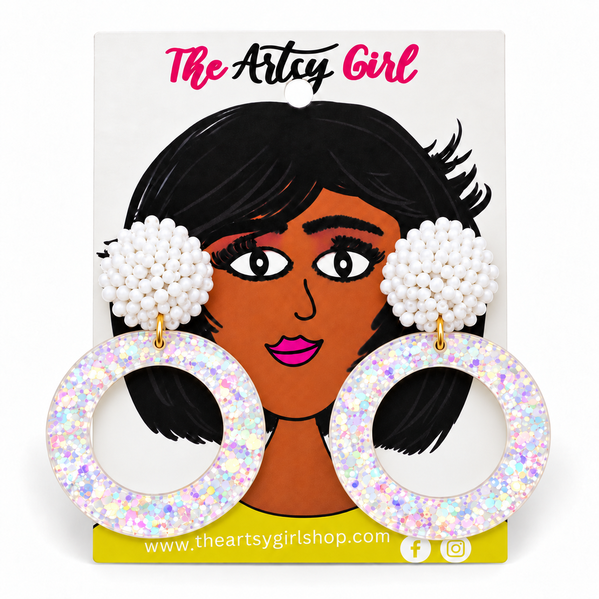 A pair of large circular hoop earrings with white pom-pom bead tops and iridescent glittery white hoops, displayed on a branded retail card that reads 'The Artcy Girl'.