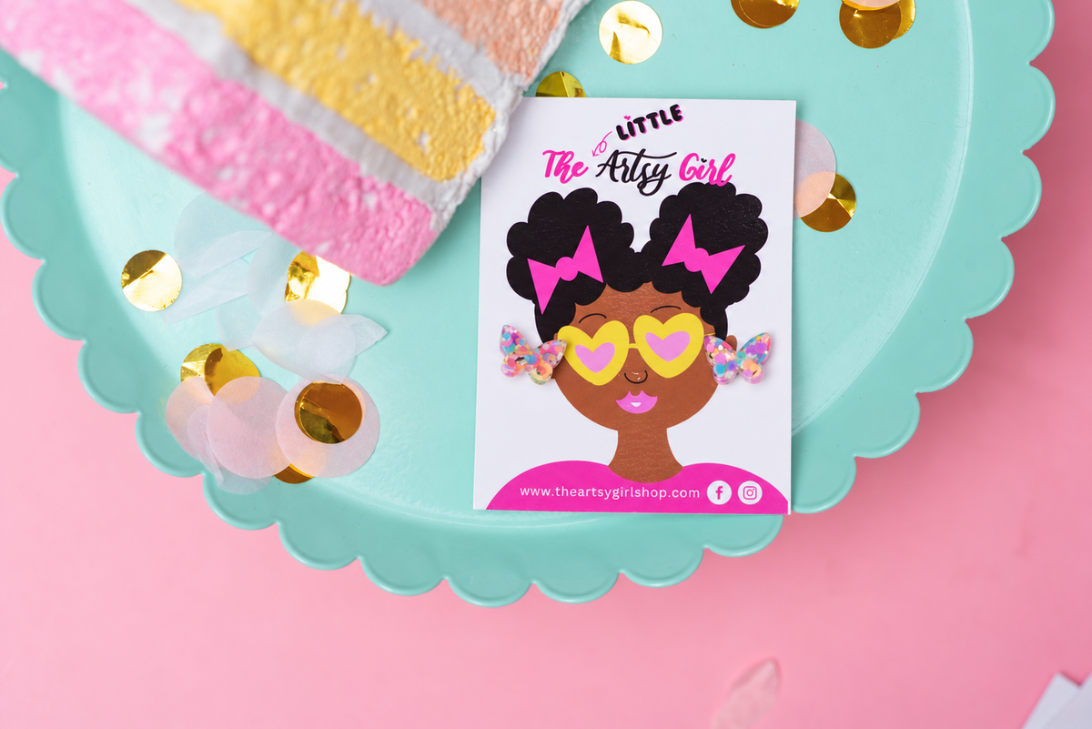 A small printed card or sticker featuring an illustrated Black woman with heart-shaped sunglasses and pink hair bows, placed on a scalloped aqua tray with gold confetti on a pink background. The card reads 'Little The Arts Girl' and includes a website at the bottom.