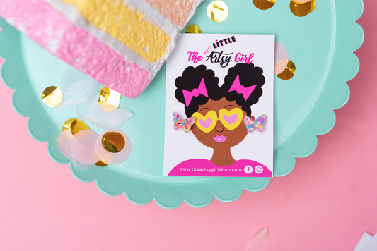 A small printed card or sticker featuring an illustrated Black woman with heart-shaped sunglasses and pink hair bows, placed on a scalloped aqua tray with gold confetti on a pink background. The card reads 'Little The Arts Girl' and includes a website at the bottom.