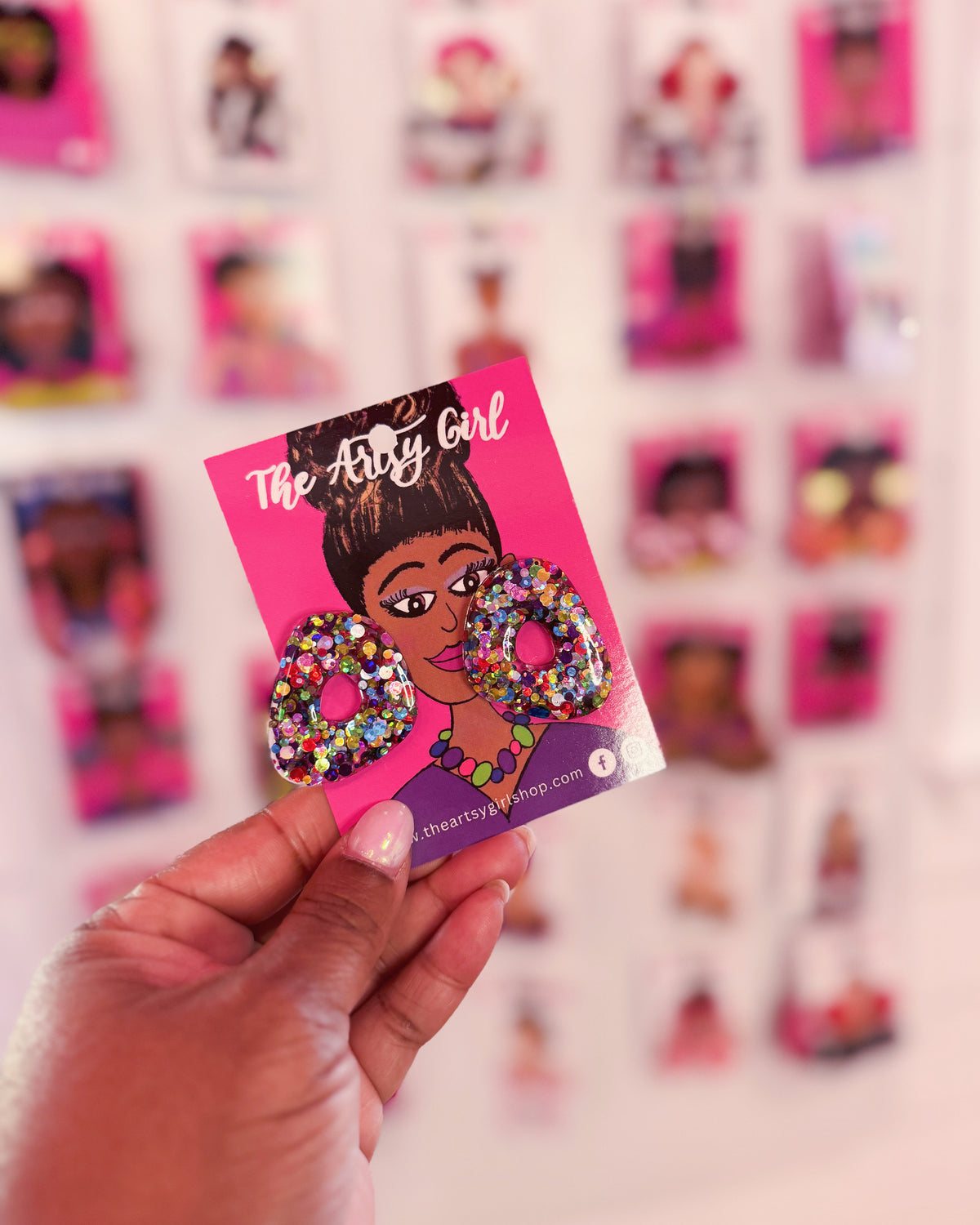 A pair of multicolored glittery earrings displayed on a pink card, with the text 'The Artsy Girl' and 'Glitz Therapy' on the earrings' description.