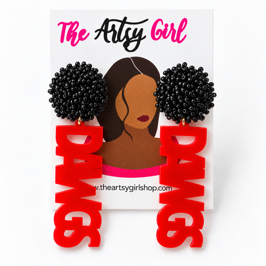 A pair of statement earrings displayed on a white card labeled 'The Artsy Girl.' The earrings have black beaded circular tops and long red letter-shaped drops spelling 'DAWGS.