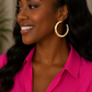 A smiling woman with long dark wavy hair wears a bright pink collared shirt and a large polished gold hoop earring, shown in a close-up portrait.